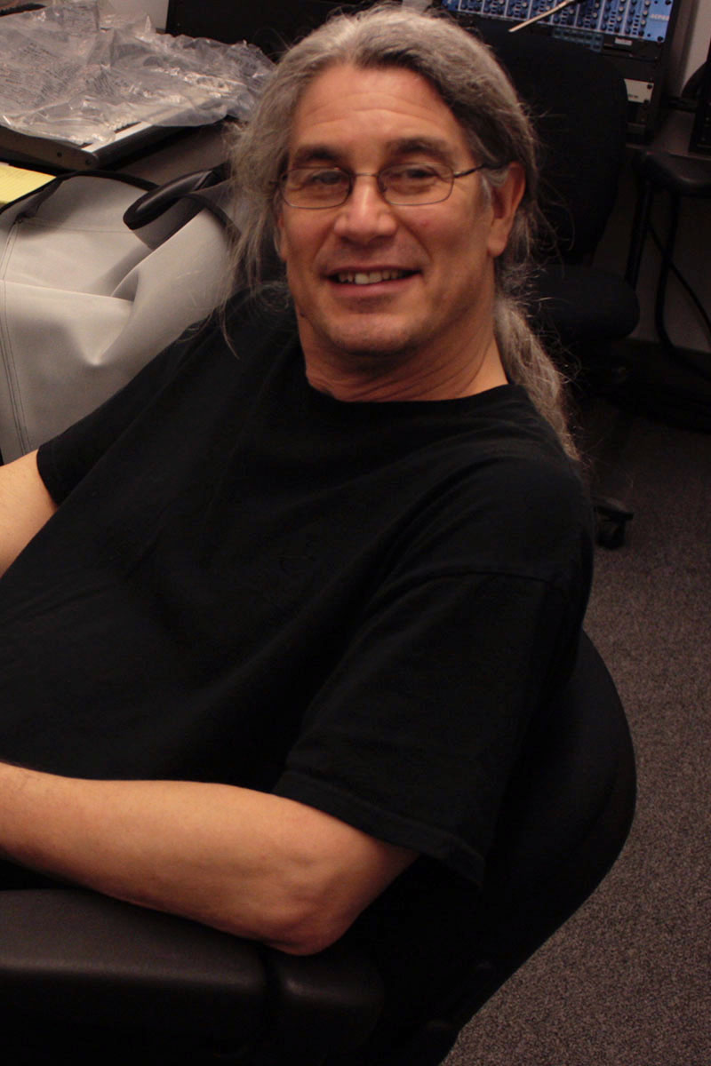 A person with long gray hair, glasses, and a black shirt smiles at the viewer.